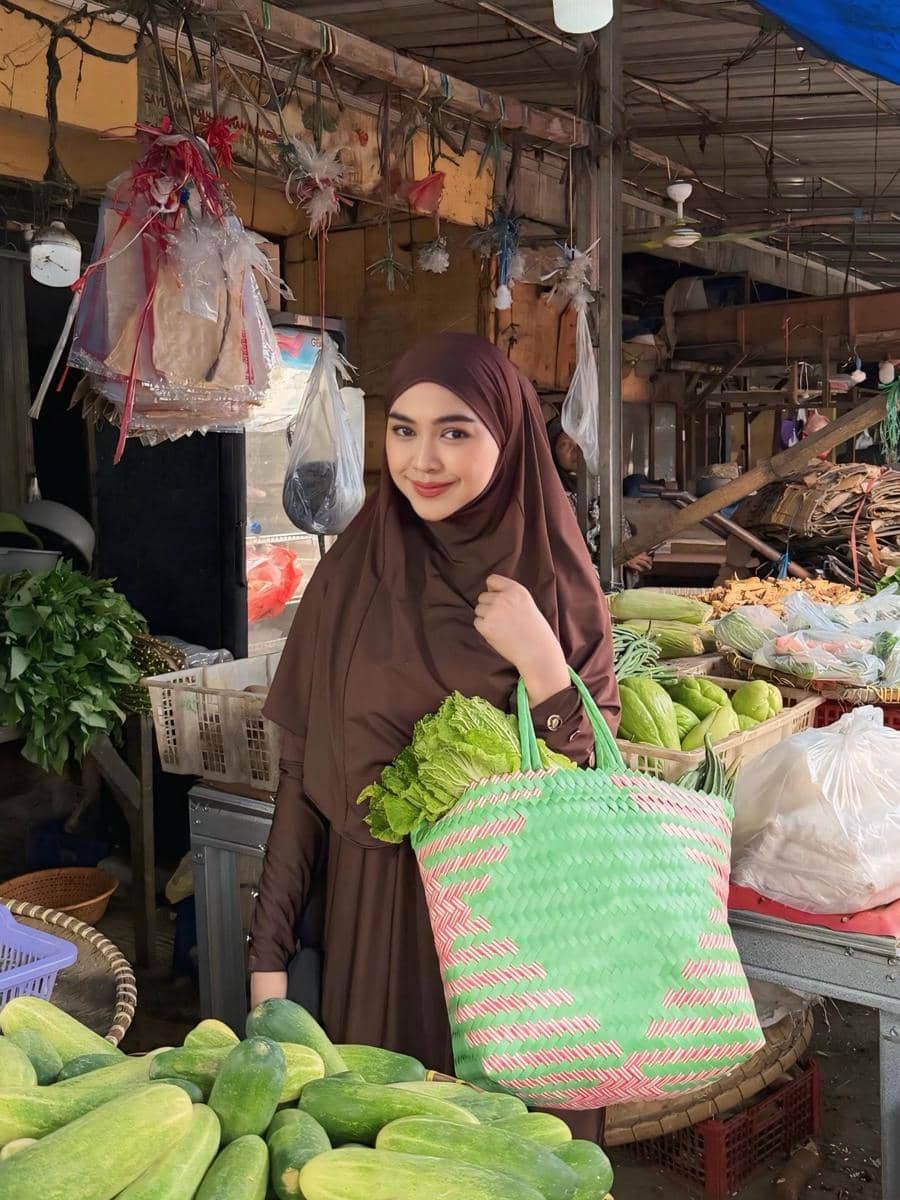Seorang wanita berhijab cokelat tersenyum sambil membawa tas anyaman berisi sayuran di pasar tradisional dengan berbagai hasil bumi.