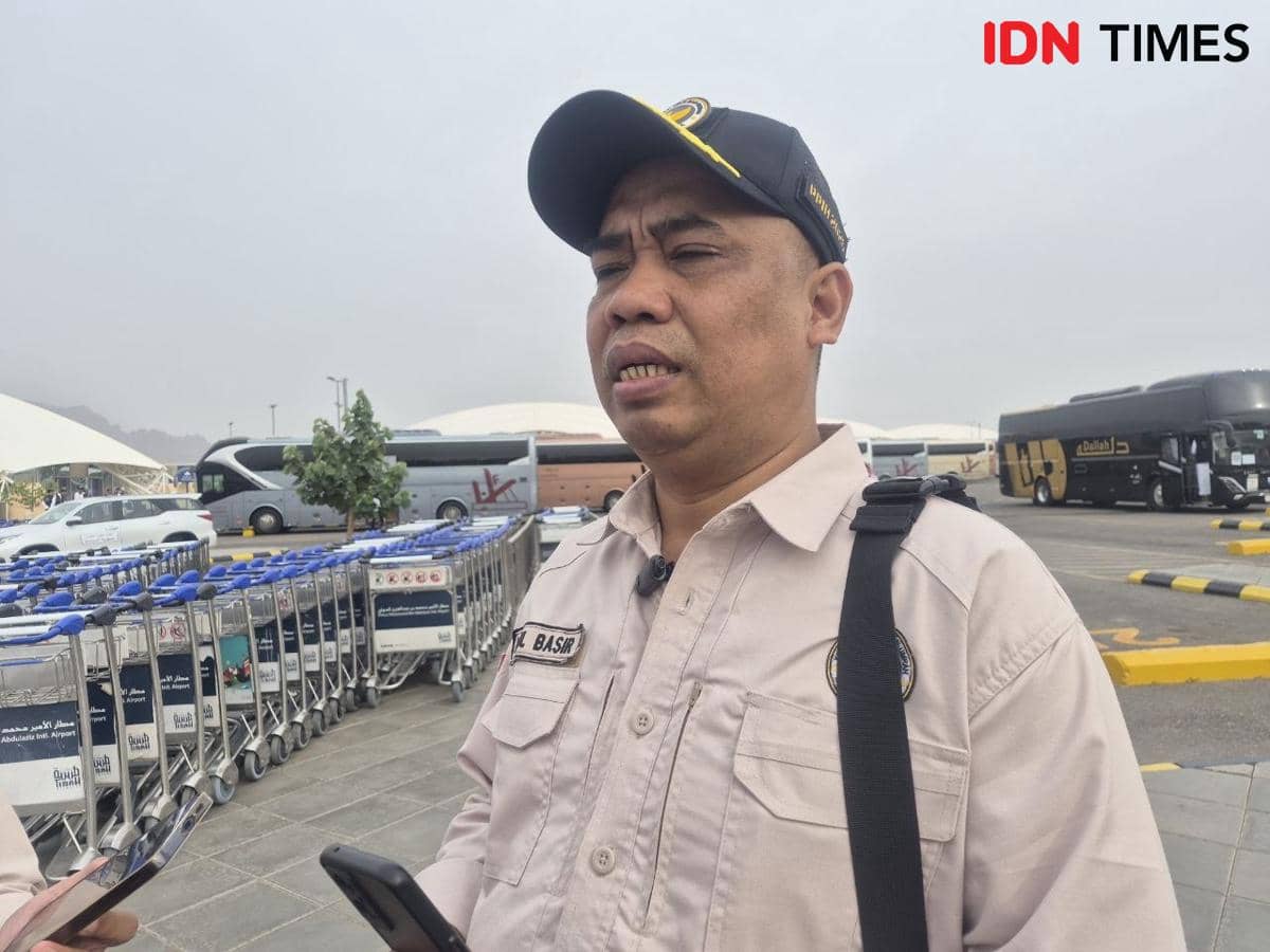 Seorang petugas berpakaian seragam krem dengan topi hitam memberikan keterangan kepada wartawan di area bandara dengan latar troli dan bus.