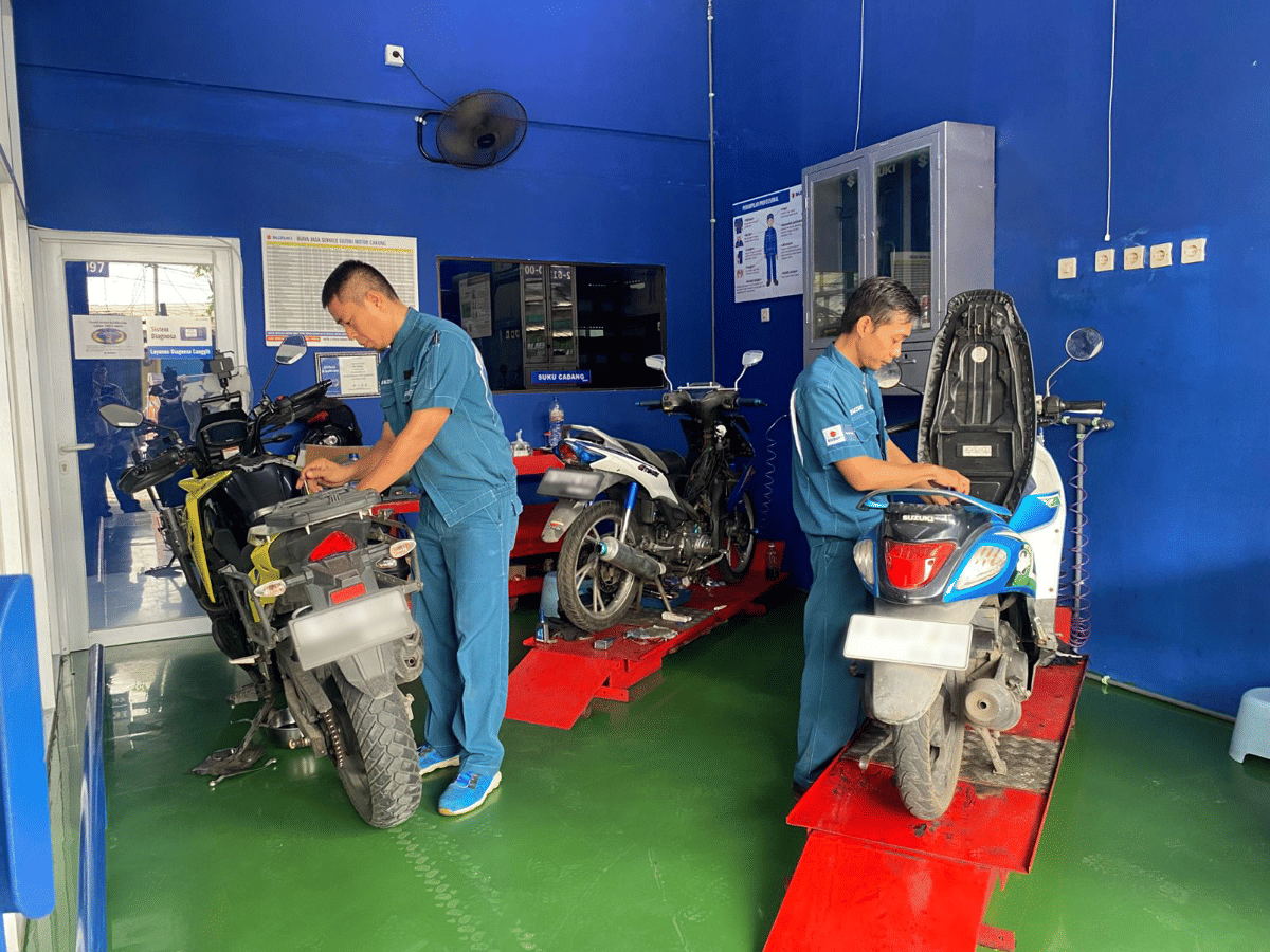 Dua mekanik bengkel sedang melakukan servis pada sepeda motor di ruang servis dengan dinding biru dan lantai hijau.