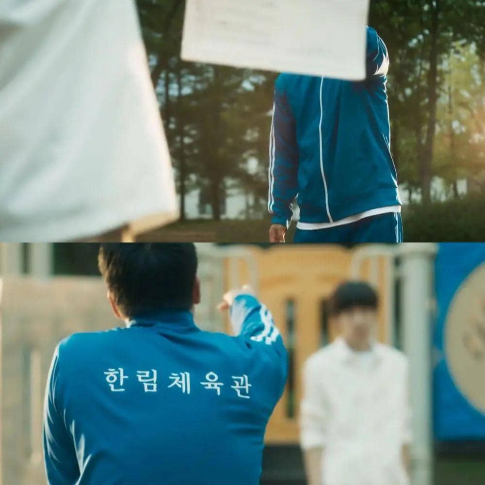 Seorang pria mengenakan jaket olahraga biru dengan tulisan Korea di punggungnya sedang berbicara dengan seseorang di luar ruangan.