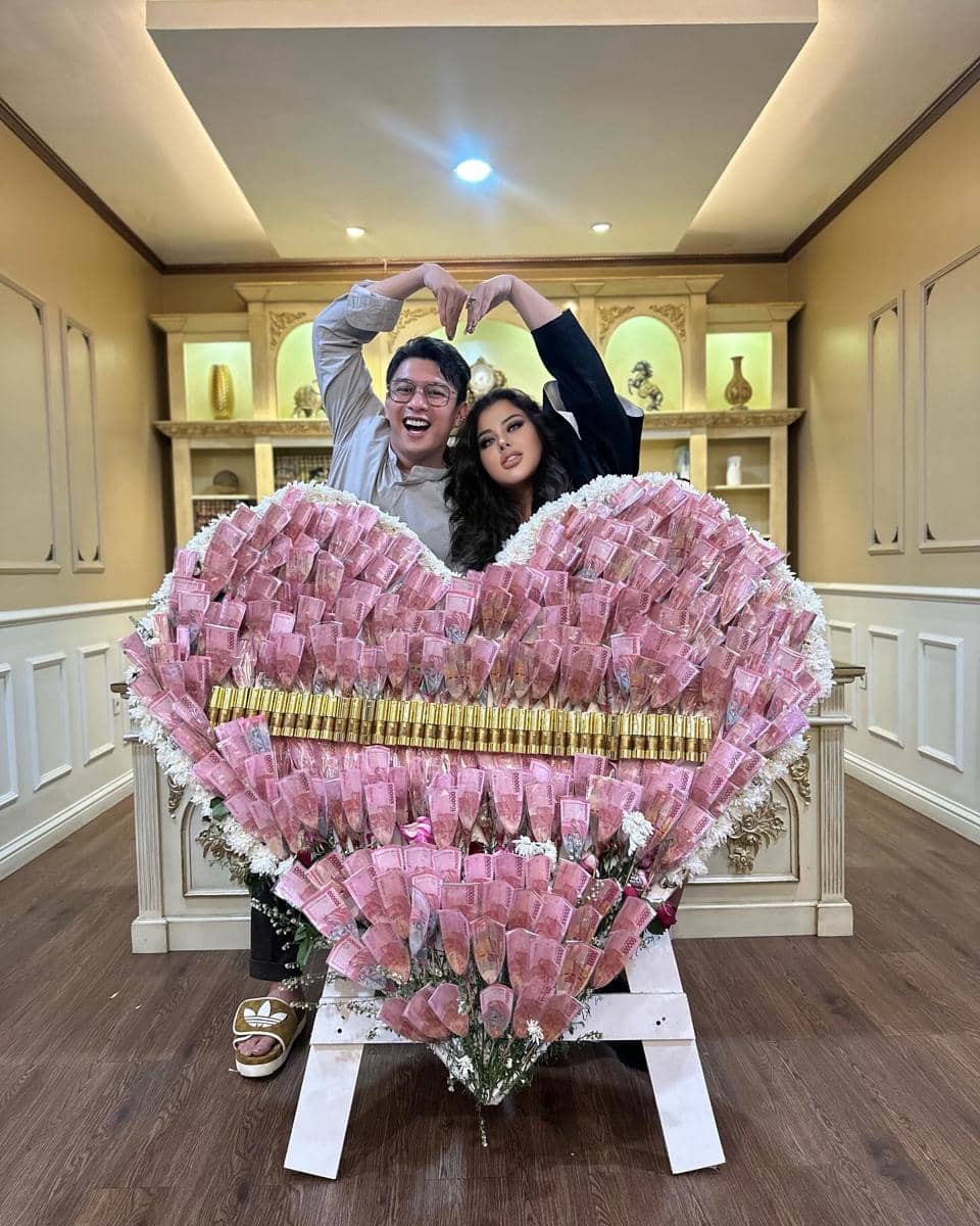 Sepasang suami istri berpose membentuk hati dengan tangan di belakang karangan bunga berbentuk hati dari uang kertas merah muda.