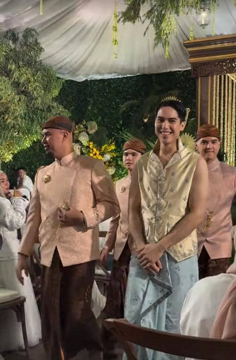 Seorang pria mengenakan busana adat Jawa berwarna krem dan biru muda tersenyum di acara pengajian dan siraman dengan dekorasi hijau dan bunga kuning.