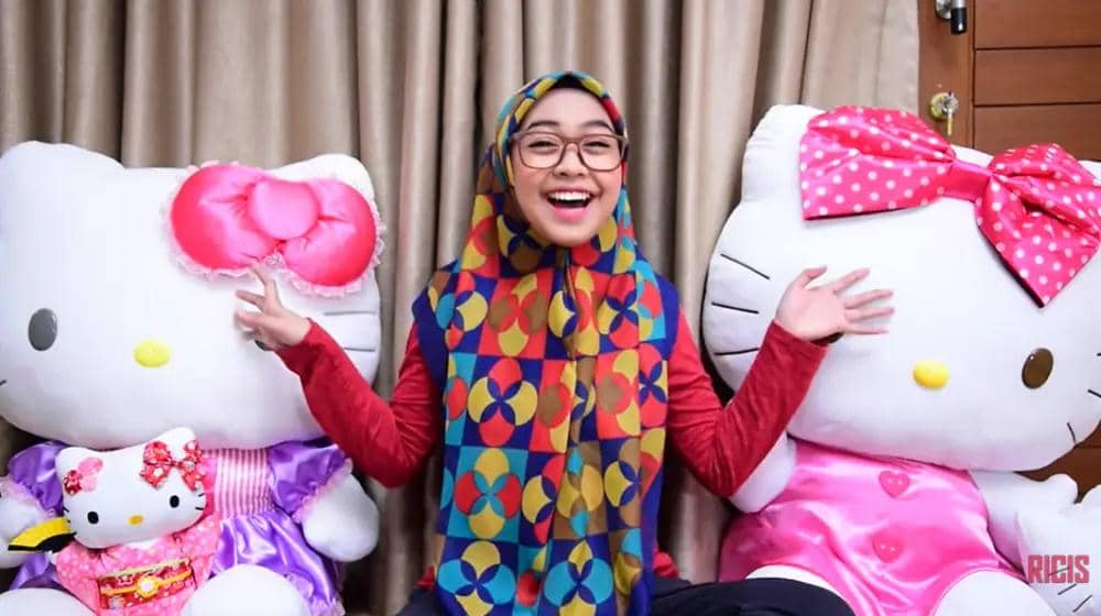 Seorang perempuan berhijab bermotif warna-warni tersenyum lebar sambil duduk di antara dua boneka besar Hello Kitty bergaun warna cerah.