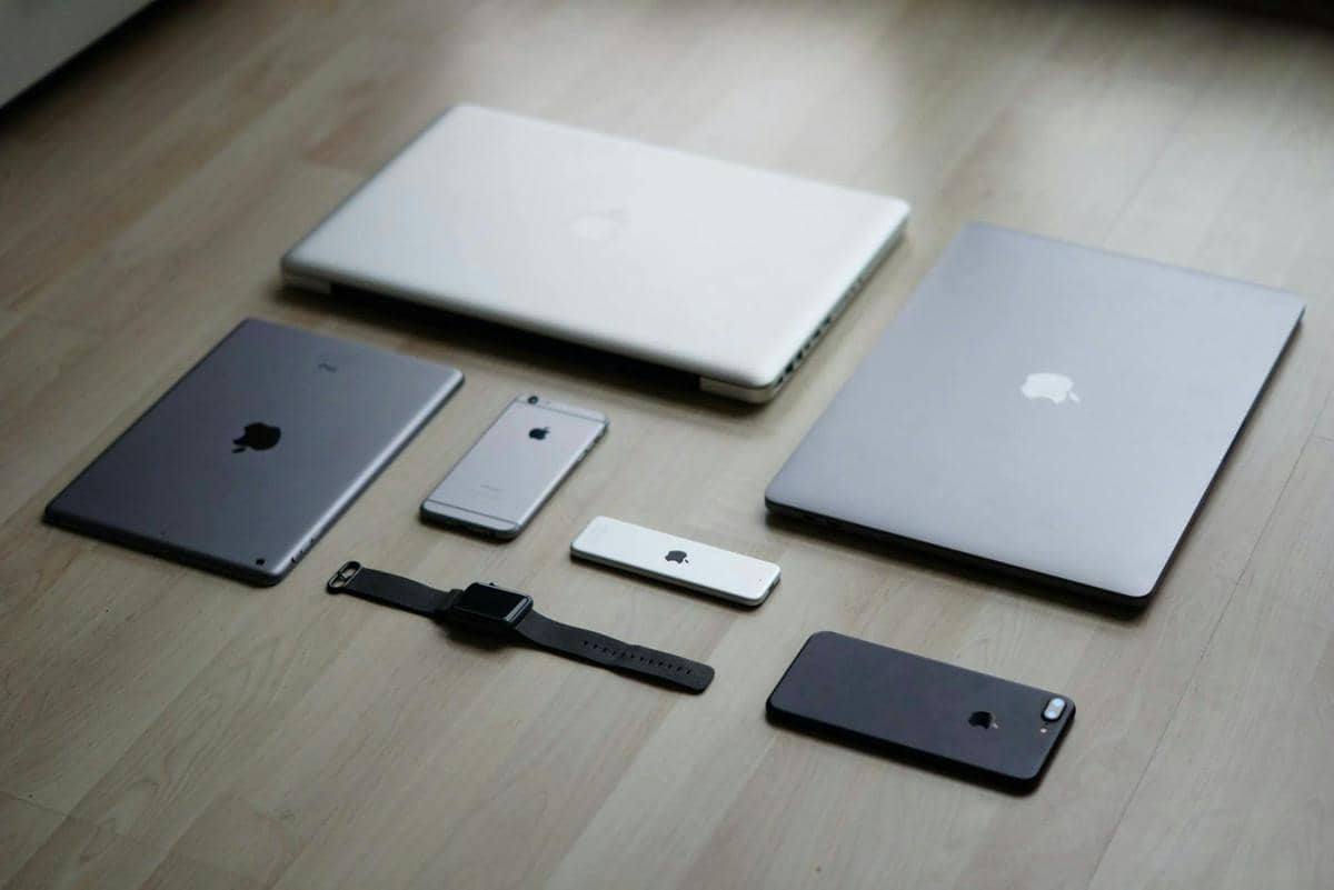 Beberapa perangkat Apple seperti MacBook, iPad, iPhone, dan Apple Watch tersusun rapi di atas lantai kayu berwarna terang.