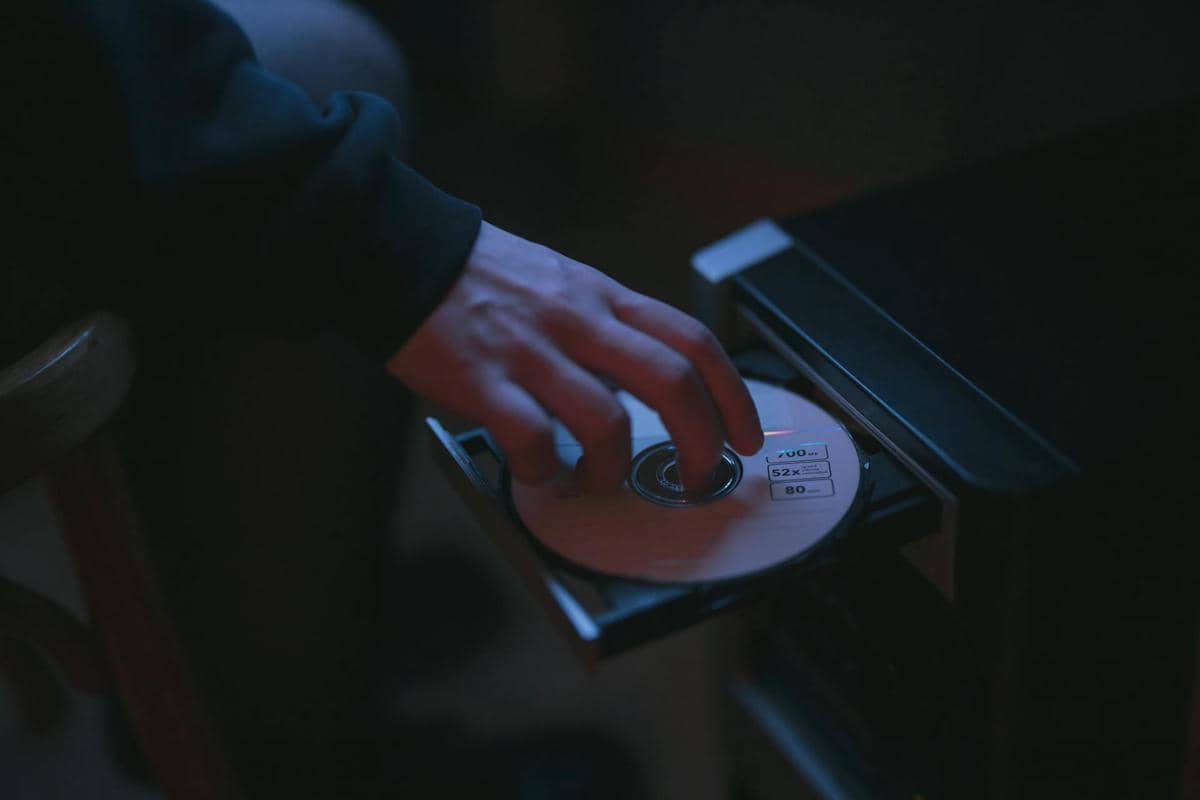 Tangan seseorang memasukkan cakram CD ke dalam pemutar atau komputer di ruangan dengan pencahayaan redup berwarna biru.