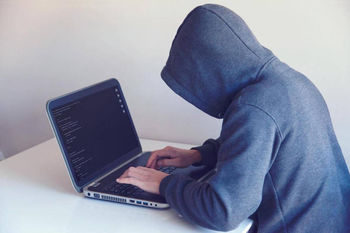 Seseorang mengenakan hoodie abu-abu sedang mengetik di laptop dengan layar menampilkan kode di atas meja putih.