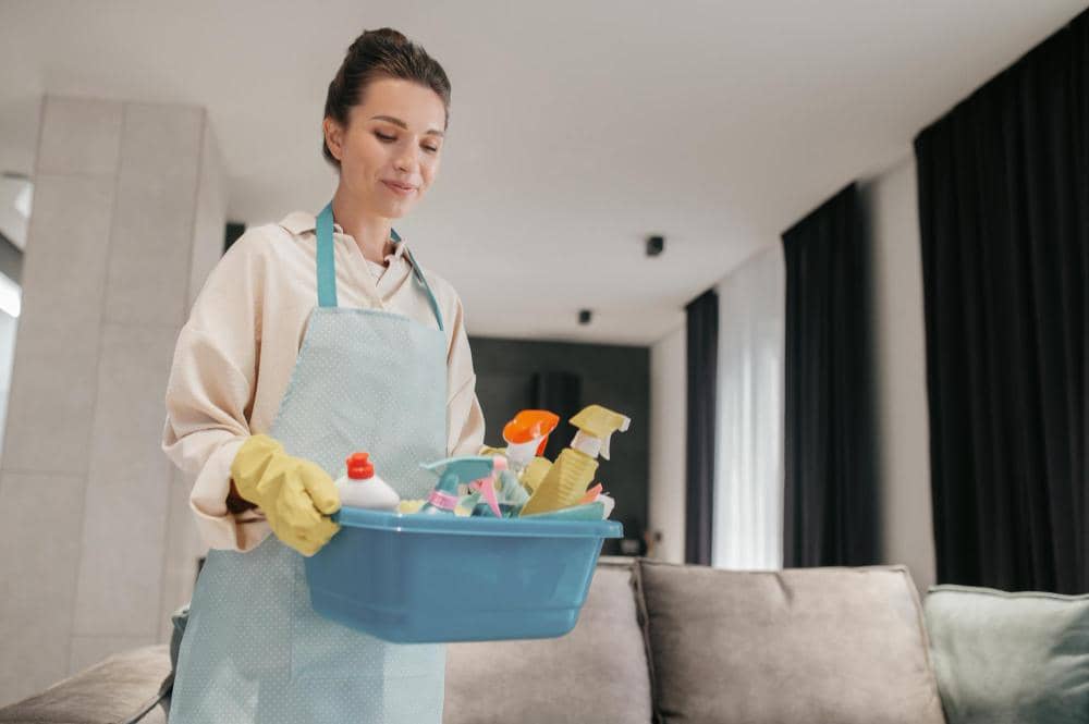 Seorang pekerja rumah tangga mengenakan celemek dan sarung tangan membawa wadah berisi berbagai alat serta cairan pembersih di ruang tamu.