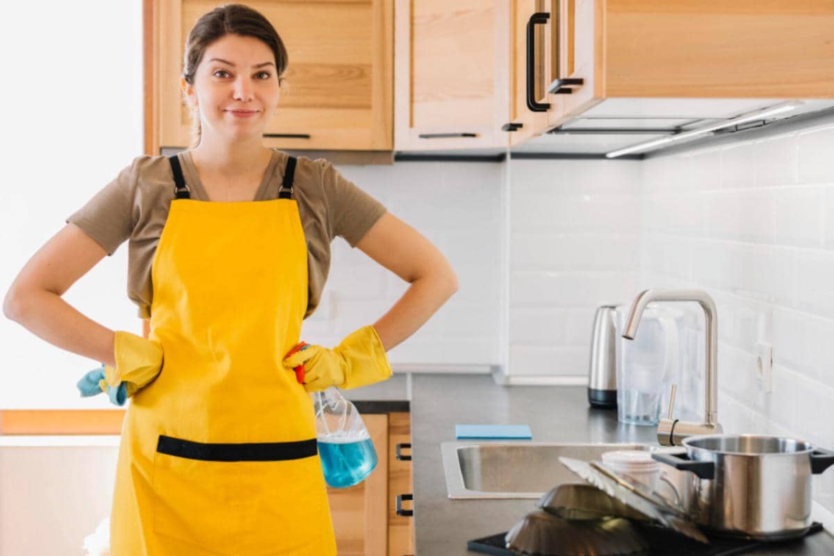 Seorang pekerja rumah tangga mengenakan celemek kuning dan sarung tangan karet sedang berdiri di dapur bersih dengan peralatan memasak di meja.