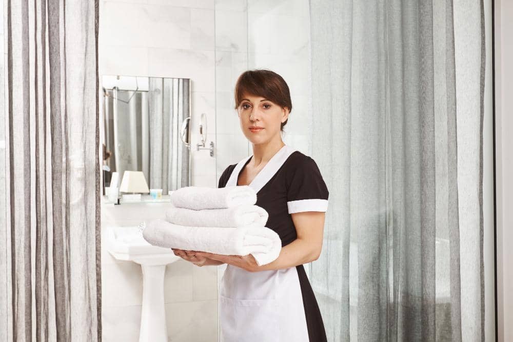 Seorang pekerja rumah tangga mengenakan seragam hitam putih sambil memegang tumpukan handuk bersih di dalam kamar mandi modern.