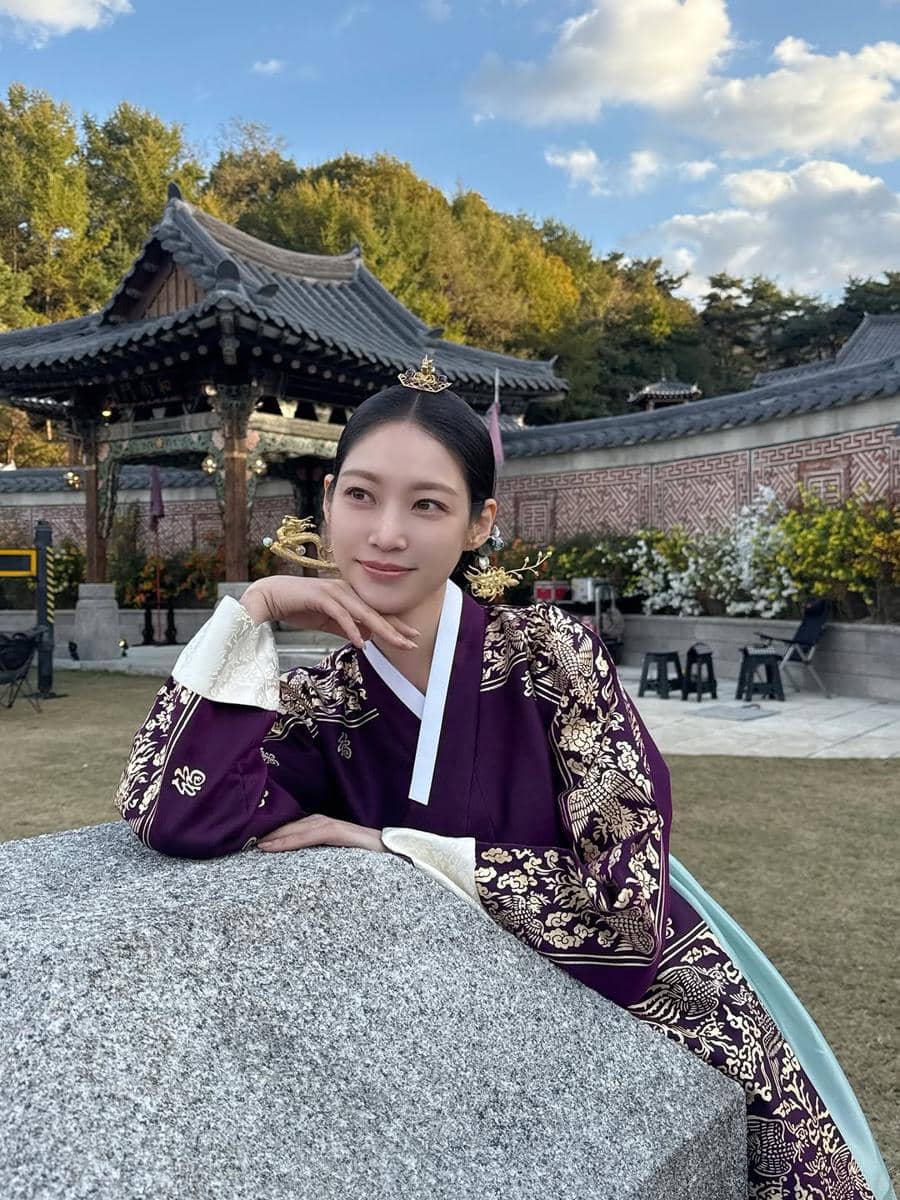 Seorang wanita mengenakan hanbok ungu berhias motif emas berpose di halaman tradisional Korea dengan latar paviliun dan pepohonan.