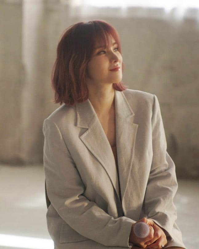 Seorang perempuan berambut pendek berwarna merah duduk sambil memegang mikrofon, mengenakan blazer abu-abu di ruangan bercahaya lembut.