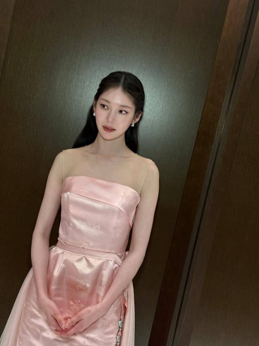 Seorang wanita berambut panjang mengenakan gaun satin berwarna merah muda lembut dengan detail sederhana berdiri di depan dinding kayu.