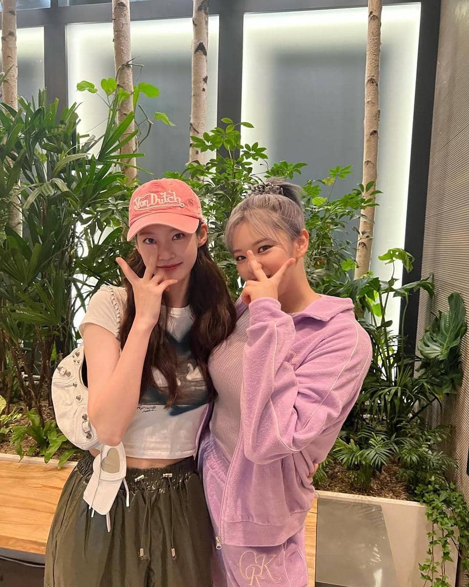 Dua wanita berpose bersama di dalam ruangan dengan latar tanaman hijau, salah satunya mengenakan jaket ungu dan lainnya topi merah muda.