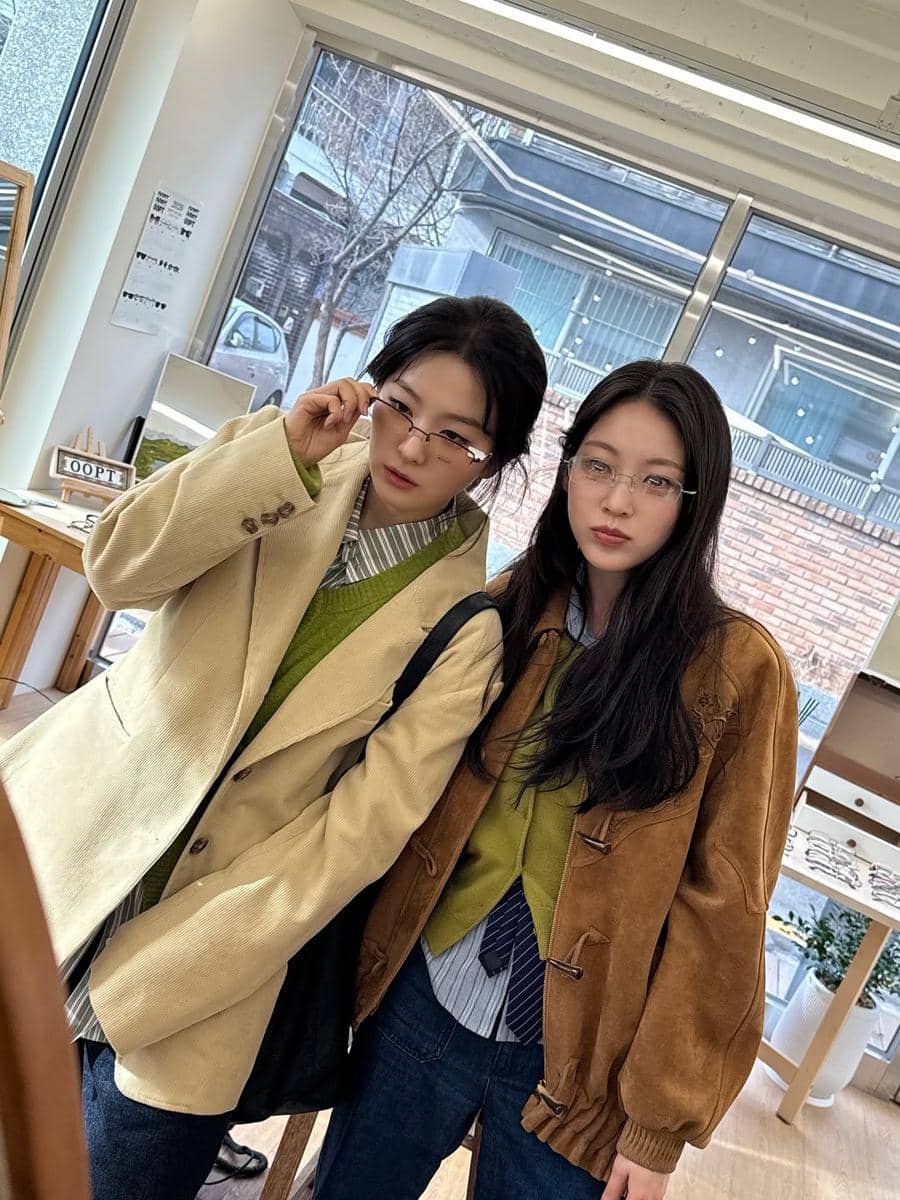 Dua wanita bergaya kasual mengenakan kacamata dan jaket berwarna cokelat serta krem di dalam ruangan dengan jendela besar.