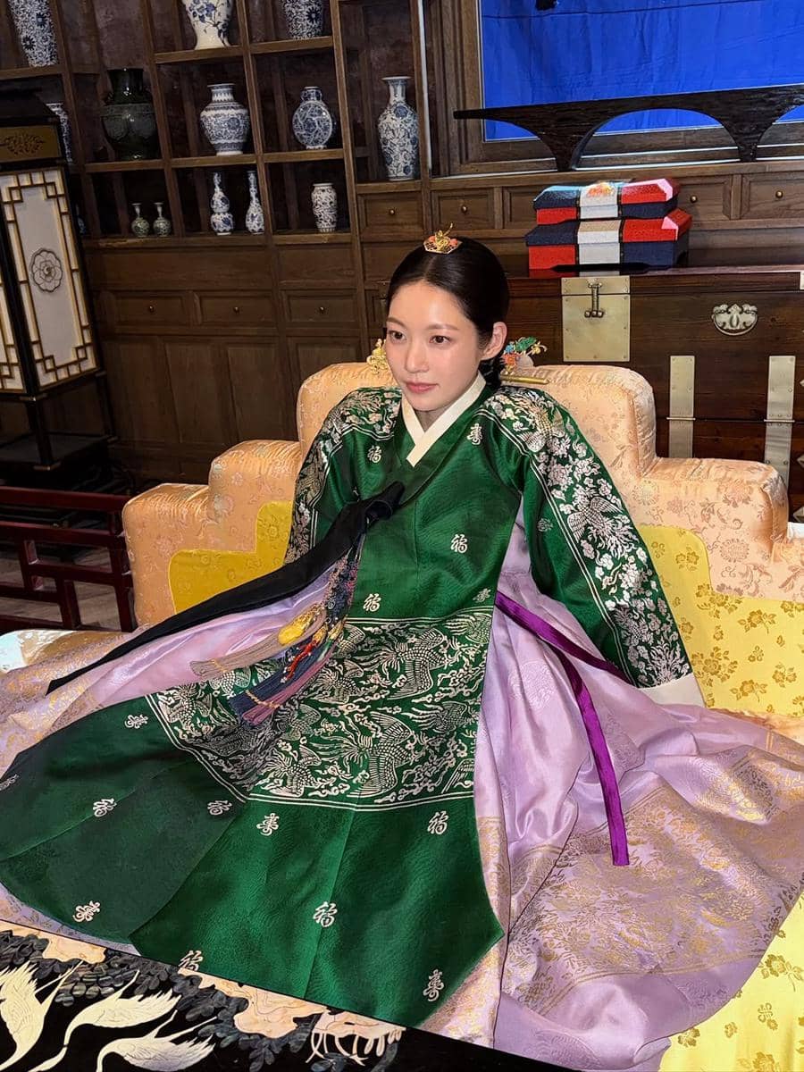 Seorang wanita mengenakan hanbok tradisional berwarna hijau dan ungu duduk di ruangan bergaya klasik dengan rak berisi guci porselen di belakangnya.