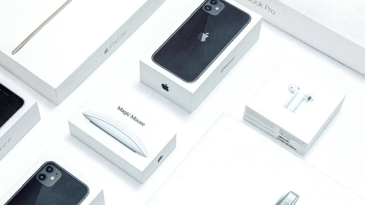Kotak kemasan berbagai produk Apple seperti iPhone, iPad Air, MacBook Pro, Magic Mouse, dan AirPods tersusun rapi di permukaan putih.