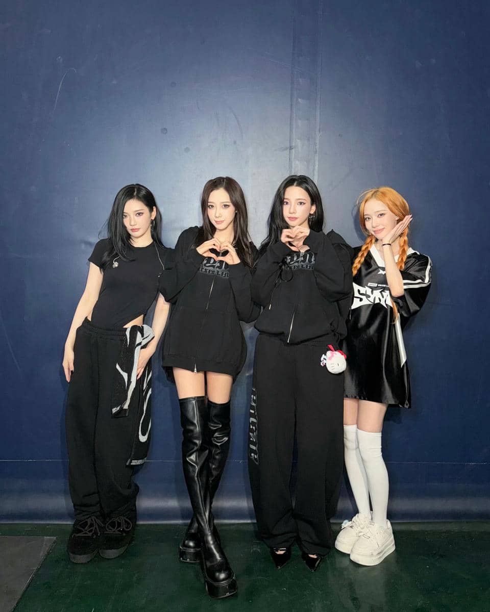 Empat anggota grup aespa berpose bersama di depan dinding biru, mengenakan busana hitam dengan gaya kasual dan modis.
