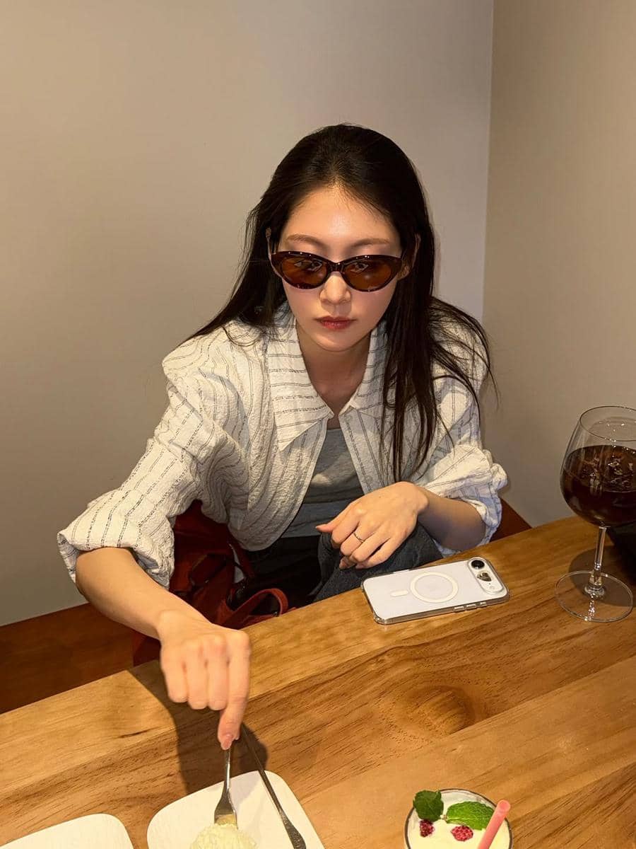 Seorang wanita mengenakan kacamata hitam duduk di meja kayu dengan makanan penutup dan minuman di depannya di sebuah kafe.