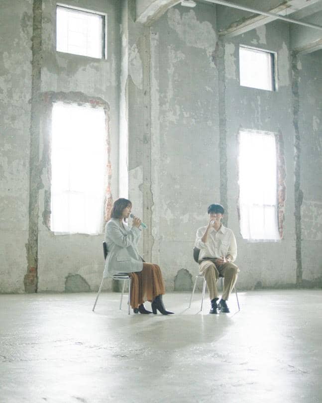 Dua orang duduk berhadapan di kursi dalam ruangan berwarna abu dengan dinding beton dan cahaya alami dari jendela besar.