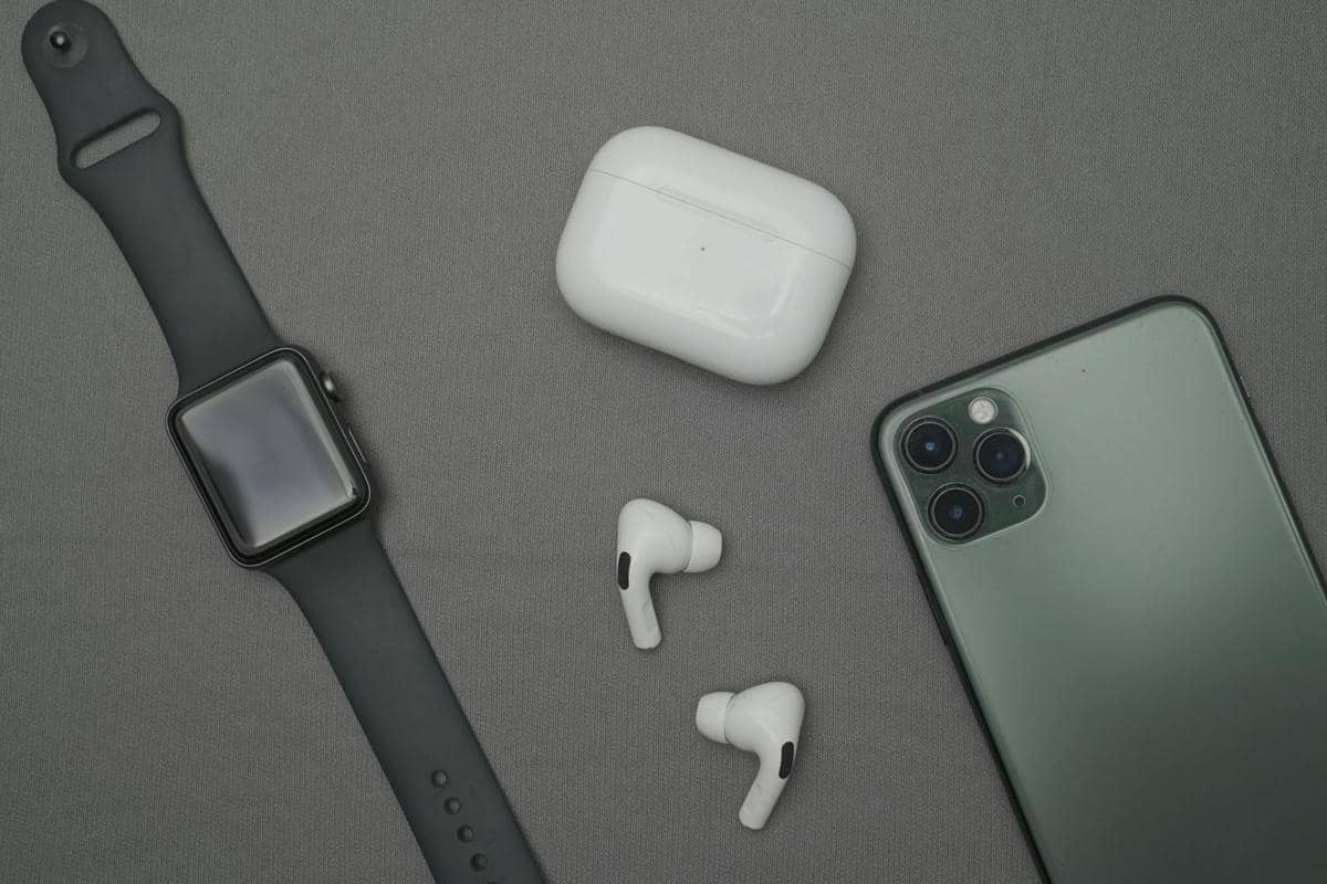 Jam tangan pintar hitam, ponsel hijau, dan sepasang earphone nirkabel putih dengan wadah pengisi daya di atas permukaan abu-abu.