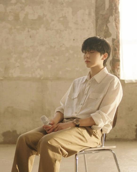 Seorang pria berambut hitam duduk di kursi sambil memegang mikrofon, mengenakan kemeja putih dan celana cokelat di ruangan bernuansa hangat.