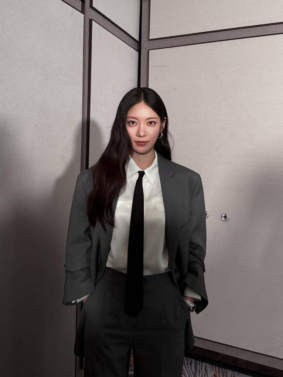 Seorang wanita berambut panjang mengenakan setelan jas abu-abu dengan kemeja putih dan dasi hitam berdiri di depan dinding panel.