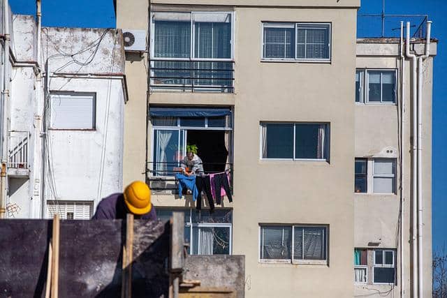 Seorang pekerja rumah tangga menggantung pakaian di balkon apartemen, dengan latar belakang gedung bertingkat dan langit cerah.