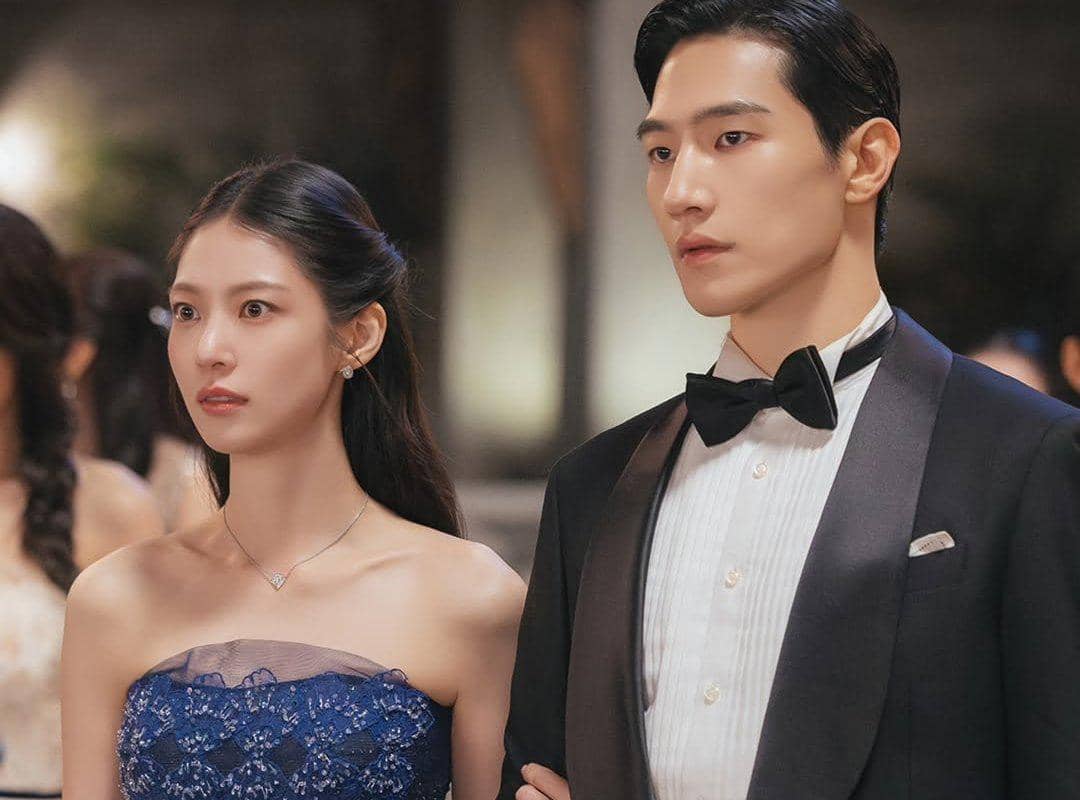 Dua pemeran drama Korea Perfect Crown mengenakan busana formal, wanita dengan gaun biru dan pria dengan setelan jas hitam serta dasi kupu-kupu.