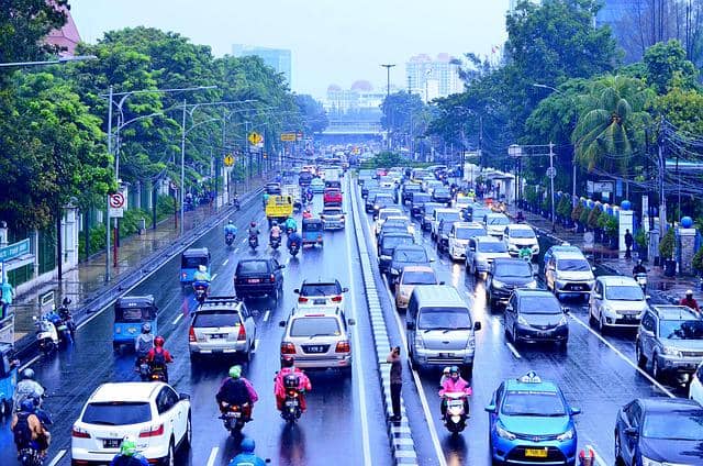 Lalu lintas padat di jalan utama Jakarta saat hari hujan dengan deretan mobil dan motor melintas di antara gedung dan pepohonan.