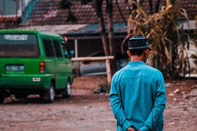 Seorang pria mengenakan baju koko biru dan peci berdiri membelakangi kamera di area luar dengan angkot hijau di latar belakang.