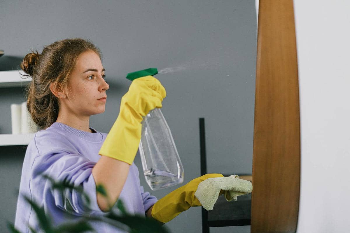 Seorang perempuan mengenakan sarung tangan karet kuning sedang menyemprotkan cairan pembersih ke cermin di dalam rumah.