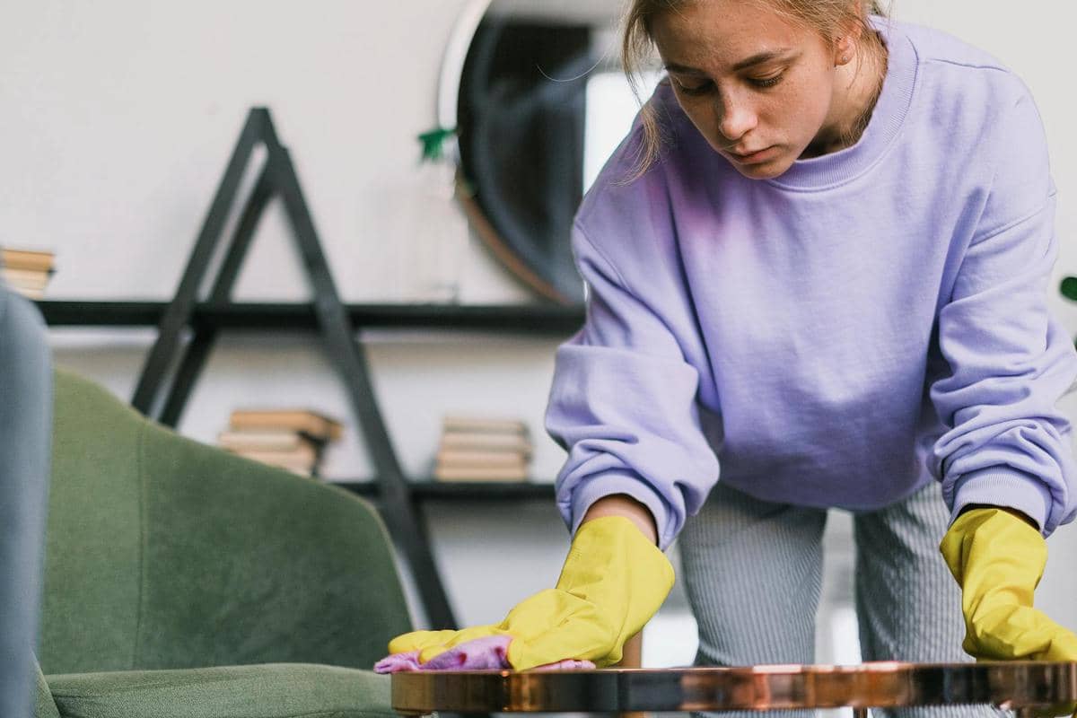 Seorang perempuan mengenakan sarung tangan karet kuning sedang membersihkan meja di ruang tamu dengan kain lap berwarna ungu.