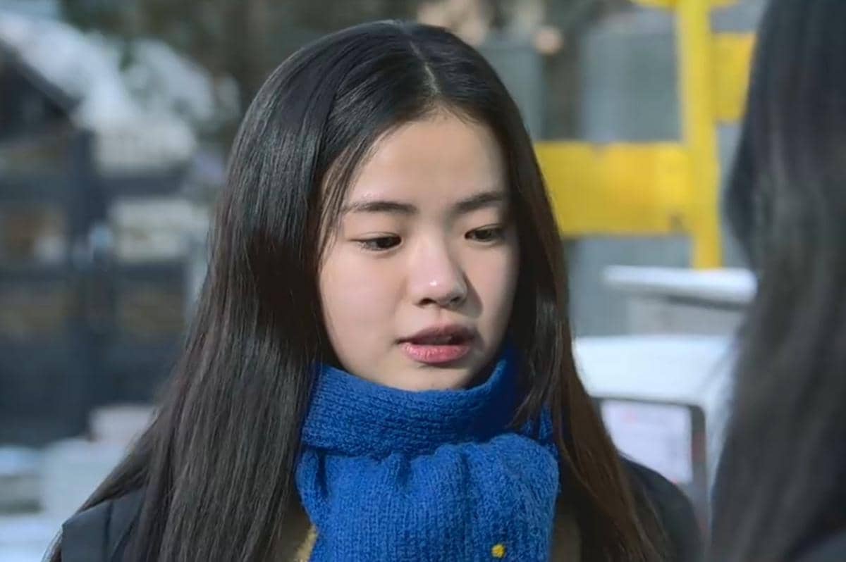 Seorang gadis berambut panjang mengenakan syal biru tebal sedang berbicara di luar ruangan dengan latar suasana dingin.