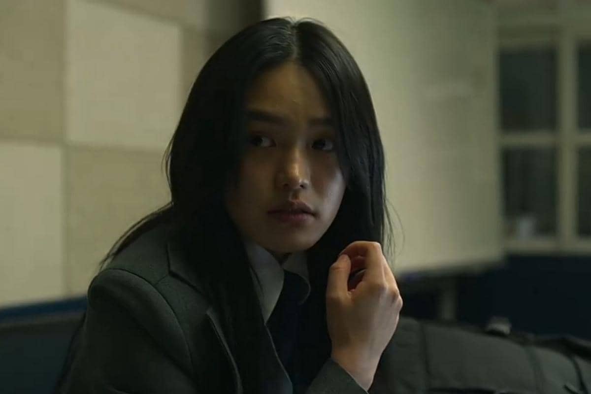 Seorang perempuan berambut panjang mengenakan seragam sekolah duduk di dalam ruangan dengan ekspresi serius dalam adegan If Wishes Could Kill.