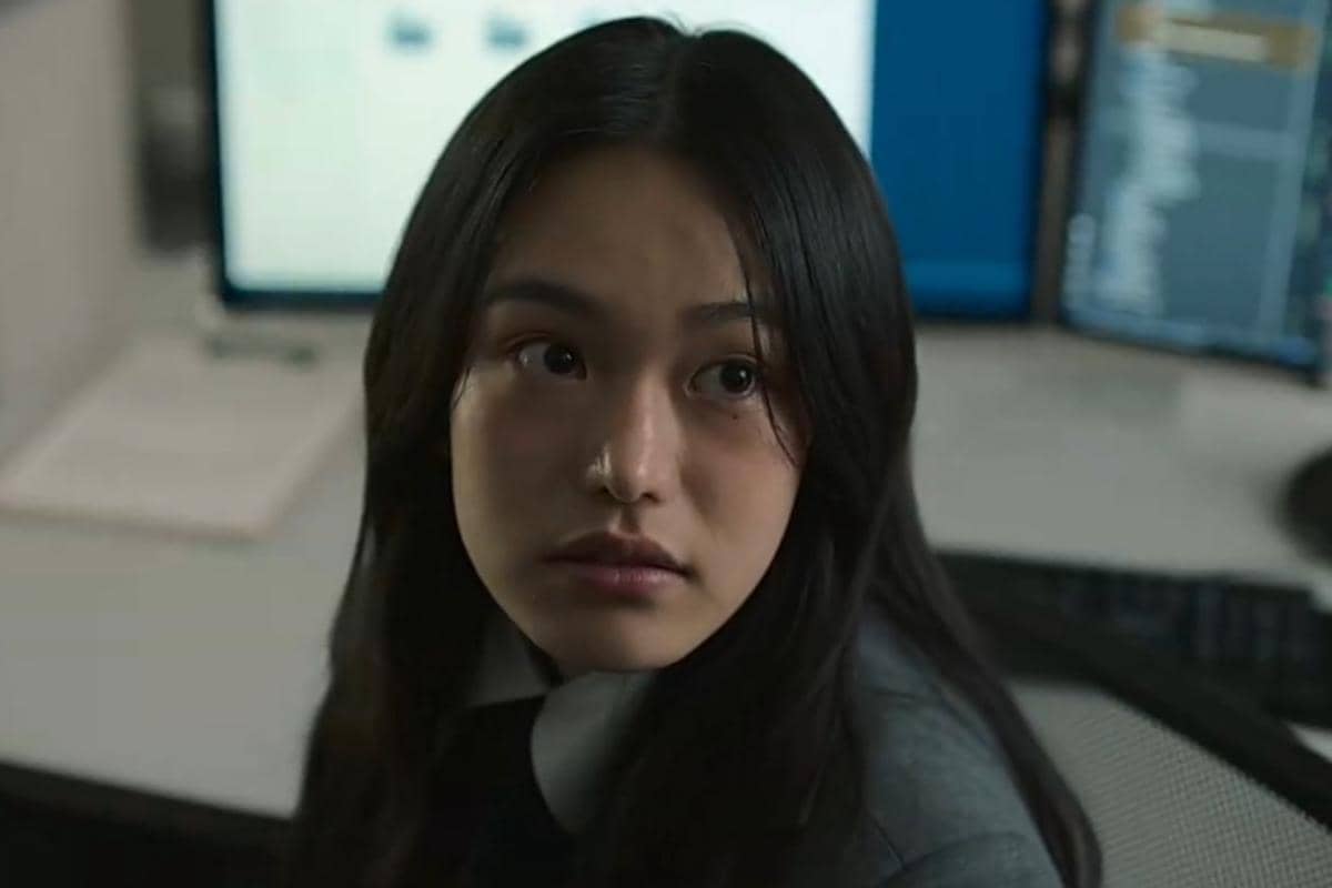 Seorang perempuan berambut panjang mengenakan pakaian formal duduk di depan komputer dengan layar menyala di latar belakang.