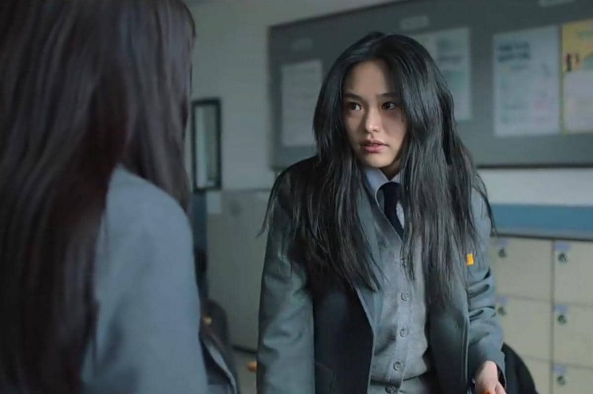 Seorang siswi berseragam abu-abu dengan rambut panjang tampak menatap serius ke arah temannya di ruang kelas sekolah.