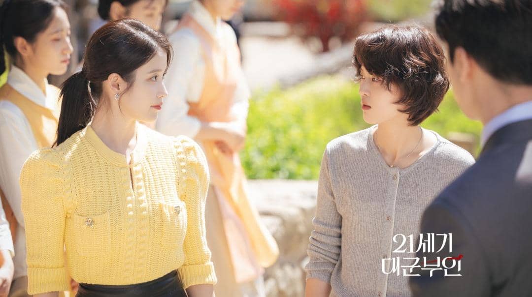 Dua wanita berdiri saling berhadapan di luar ruangan dengan latar taman, salah satunya mengenakan sweater kuning dan yang lain abu-abu.