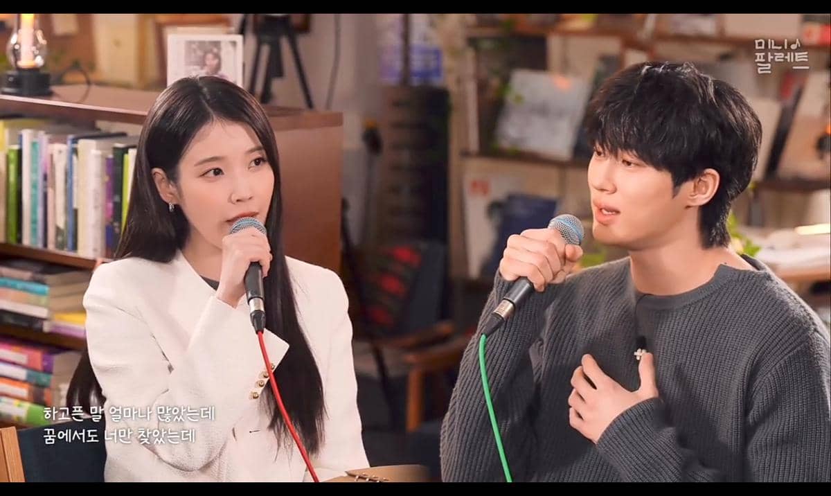 Dua penyanyi duduk berhadapan sambil memegang mikrofon dan bernyanyi bersama di sebuah ruangan dengan latar rak buku.