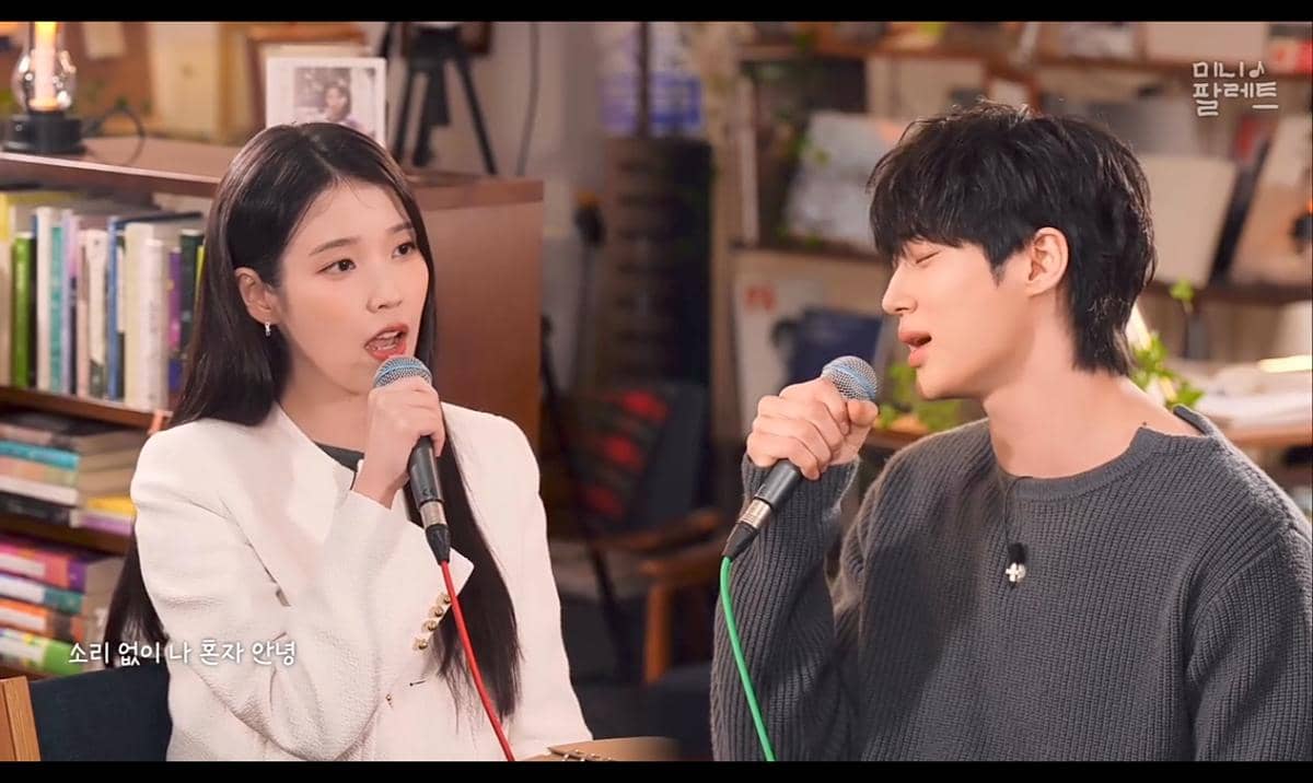 Dua penyanyi sedang berduet sambil memegang mikrofon di studio dengan latar buku dan dekorasi hangat, menampilkan suasana intim dan fokus.