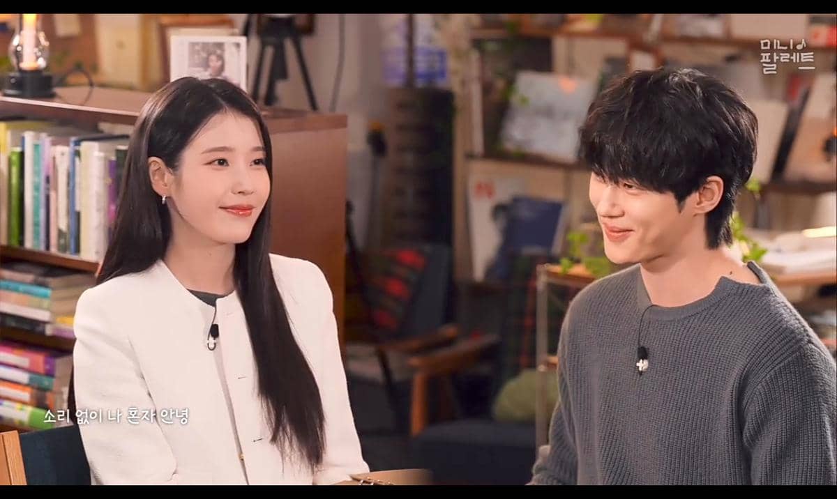 Dua orang duduk berhadapan sambil tersenyum di sebuah ruangan hangat dengan latar rak buku dan dekorasi bernuansa kayu.