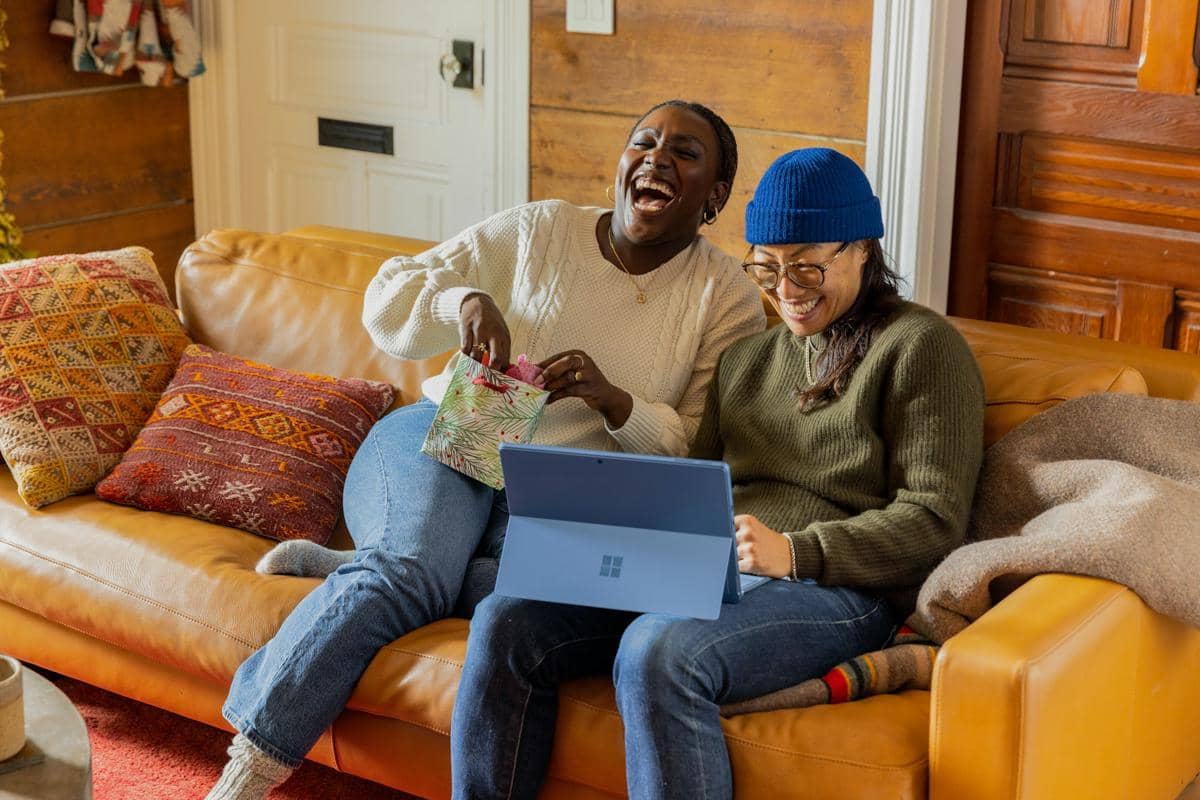 Dua orang duduk di sofa berwarna cokelat sambil tertawa bersama, satu membuka hadiah dan satu menggunakan laptop di ruang tamu hangat.
