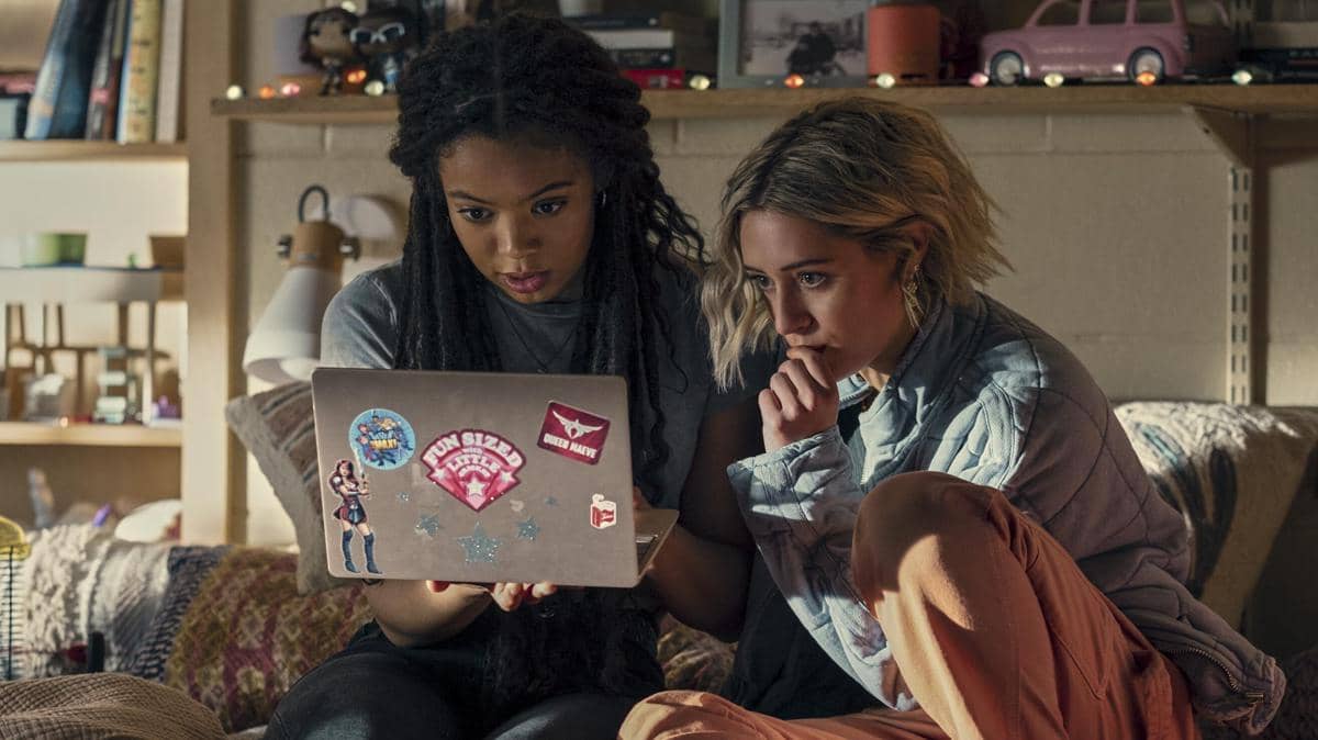 Dua perempuan muda duduk di kamar asrama sambil menatap layar laptop berstiker warna-warni dengan ekspresi serius.