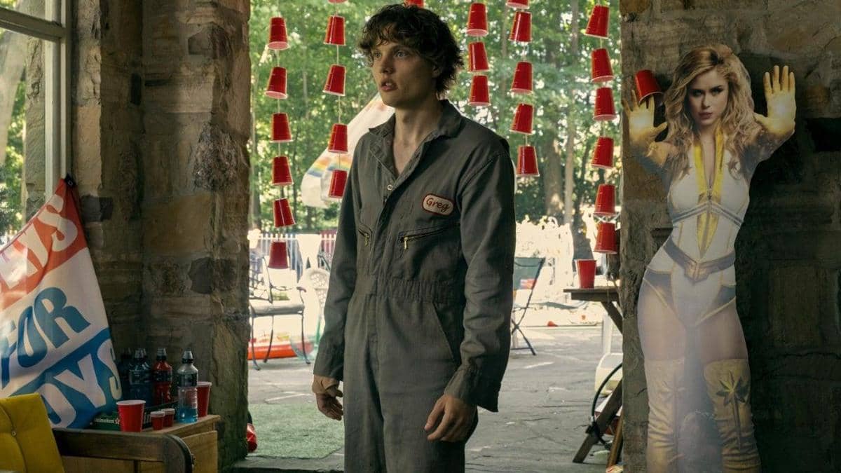 Seorang pria mengenakan jumpsuit abu-abu dengan nama 'Greg' berdiri di ruangan berhias gelas plastik merah dan poster karakter perempuan superhero.