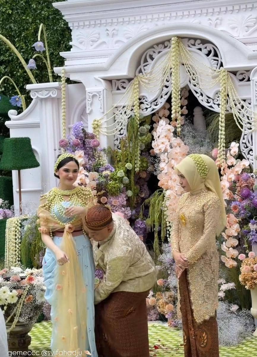 Seorang perempuan mengenakan kebaya modern warna pastel dengan hiasan kepala tradisional berdiri di acara pengajian dan siraman bernuansa bunga.