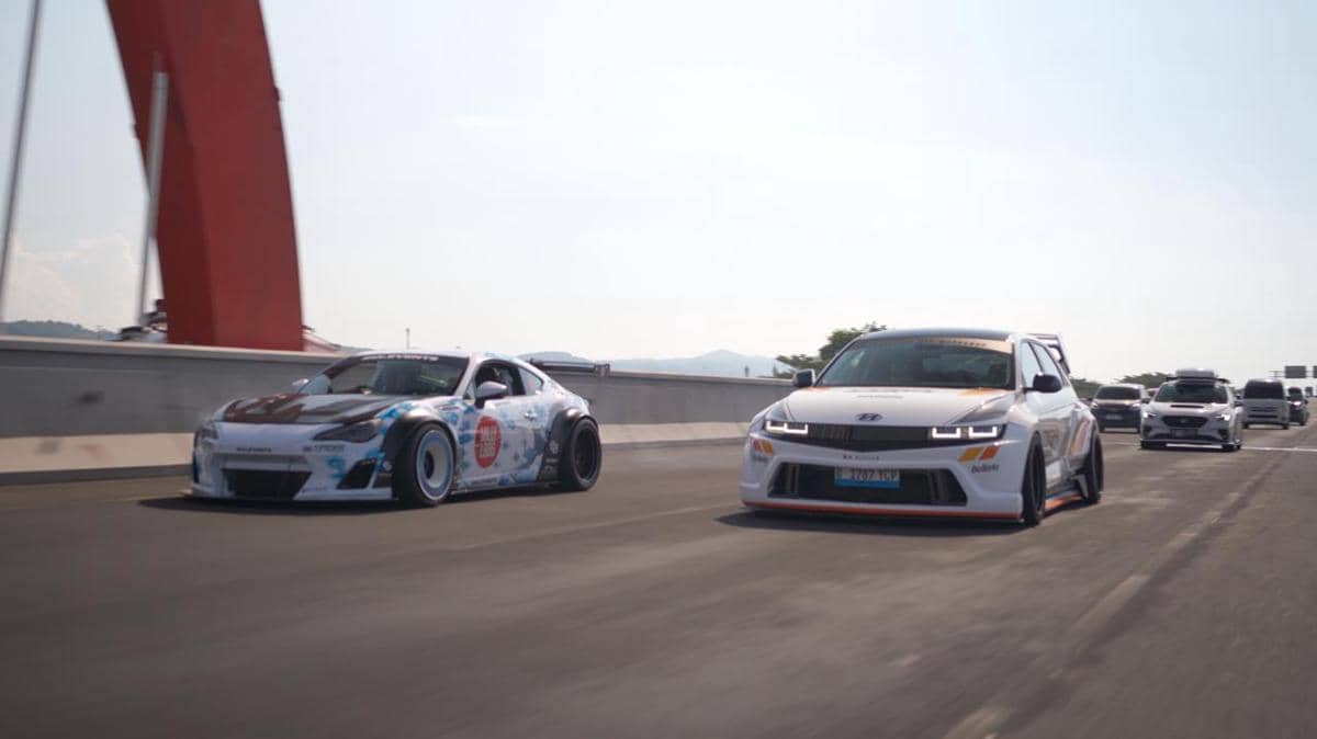 Dua mobil modifikasi melaju berdampingan di jalan raya dengan latar jembatan merah dan langit cerah di siang hari.