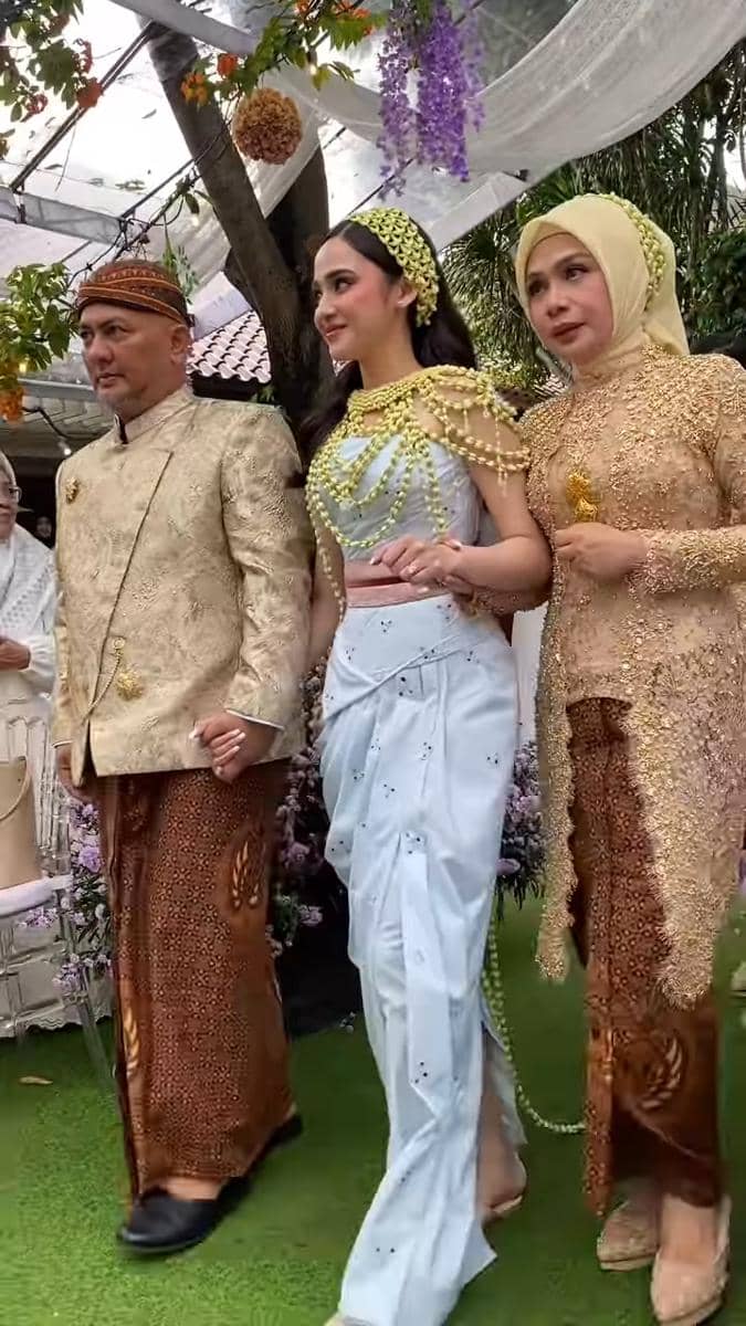 Seorang perempuan mengenakan kebaya modern berwarna biru muda dengan hiasan melati, berjalan diapit dua orang berpakaian adat Jawa.