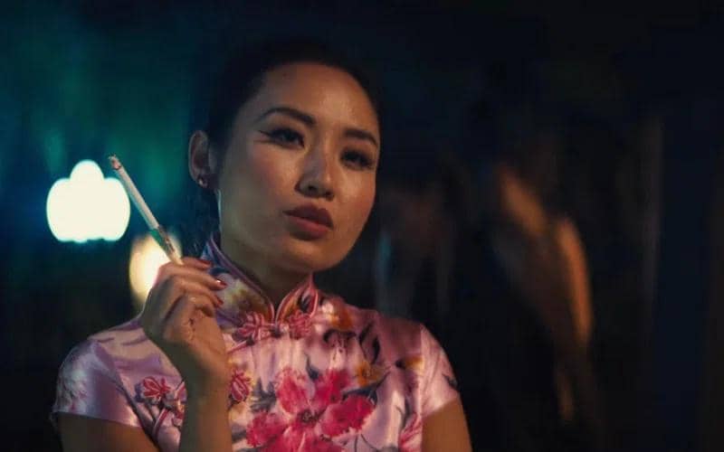 Seorang perempuan mengenakan cheongsam berwarna merah muda bermotif bunga memegang rokok di tangan dalam suasana malam yang remang.