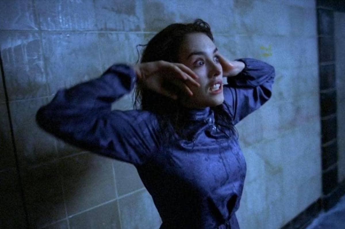 Seorang wanita dengan ekspresi ketakutan dan panik berdiri di ruangan berdinding ubin biru, mengenakan pakaian gelap yang basah.