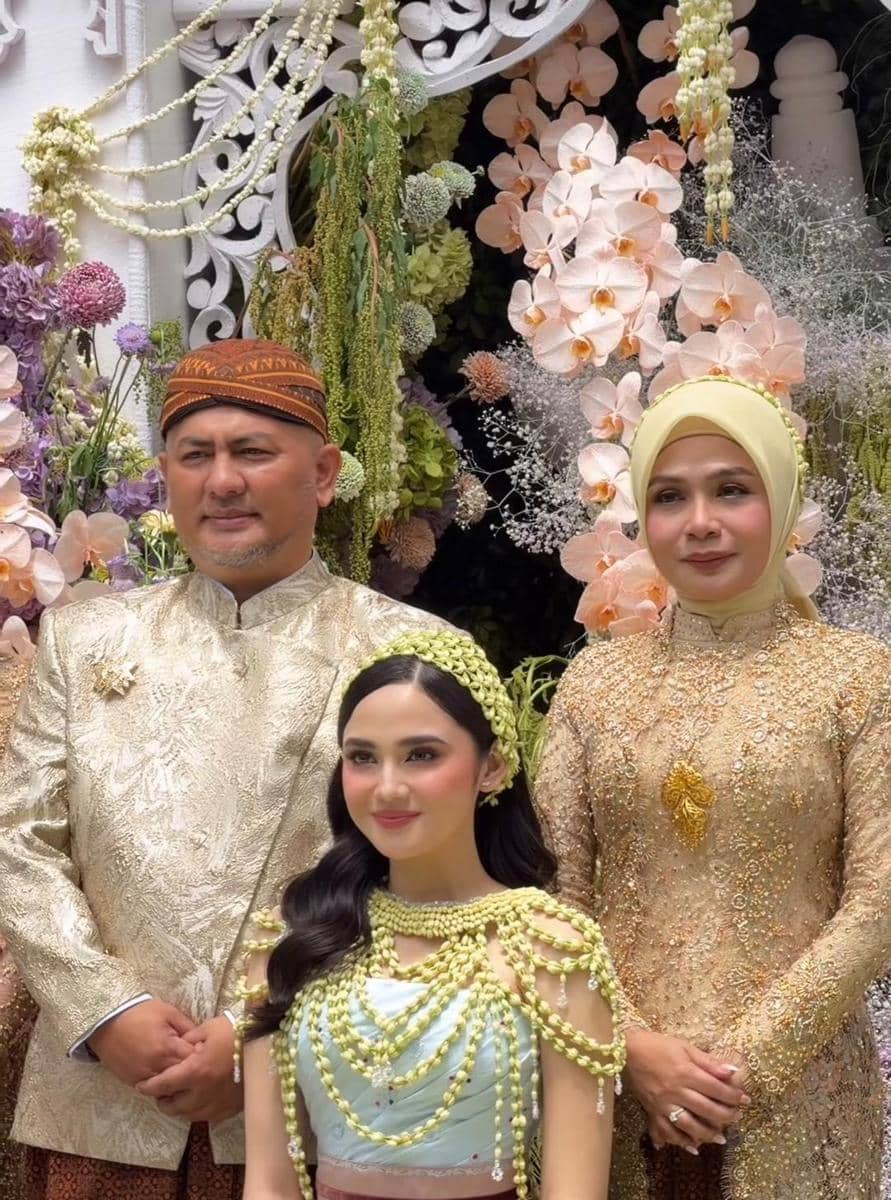 Seorang perempuan muda berbusana adat warna pastel berpose bersama dua orang dewasa berpakaian tradisional di depan dekorasi bunga.