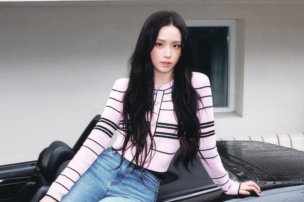 Seorang wanita berambut panjang mengenakan sweater pink bergaris hitam dan celana jeans duduk di atas mobil hitam di luar ruangan.